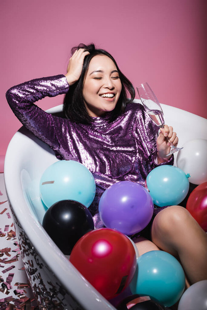 veselá asijská žena ve fialových šatech ležící ve vaně s barevnými balónky a držící sklenici šampaňského na růžové  - Fotografie, Obrázek