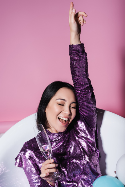 pozitivní asijská žena ve fialových šatech ležící ve vaně s barevnými balónky a drží sklenici šampaňského na růžové  - Fotografie, Obrázek