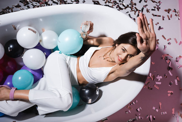 top view of positive woman with tattoo gesturing and lying in bathtub with balloons while holding glass of champagne - Foto, afbeelding