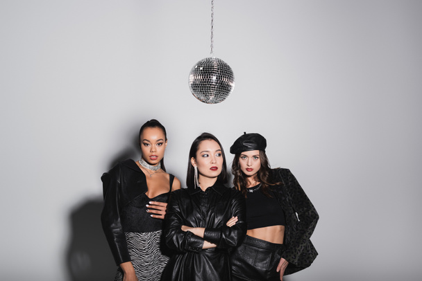 young model in leather beret posing with hand on hip near interracial women and shiny disco ball on grey  - Photo, Image