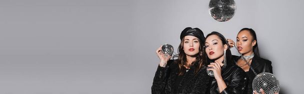 young woman in beret posing near interracial models holding disco balls on grey, banner - Foto, Imagem