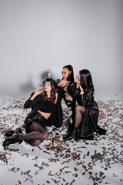 longitud completa de jóvenes modelos multiétnicos sosteniendo copas de champán mientras se sienta cerca de confeti brillante en gris - Foto, imagen