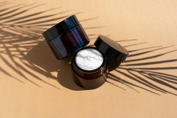 Two cosmetic creme in dark glass jars on beige background. Organic moisturizing balm with palm leaves shadow. - Фото, зображення