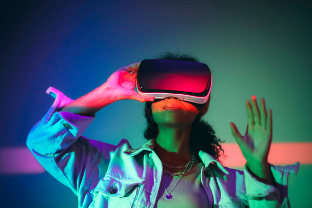 Virtual Reality. Young african american woman using VR headset goggles, interacting with cyberspace and exploring virtual world under neon light, gesturing with hand. Digital gaming and entertainment - Фото, зображення