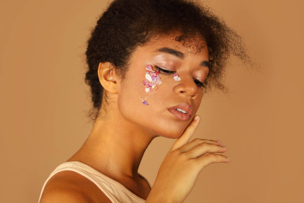 Portrait of beautiful African girl with curly hair shows creative make-up with flower petals while hand gently touches her face isolated over beige background. Natural eco cosmetics and beauty concept - Zdjęcie, obraz
