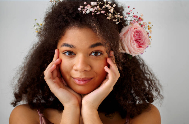 Закройте весенний портрет великолепной улыбающейся африканско-американской молодой женщины со свежим розовым цветом розы в вьющихся волосах, смотрящей в камеру, изолированной на сером фоне. Естественная красота африканских женщин - Фото, изображение