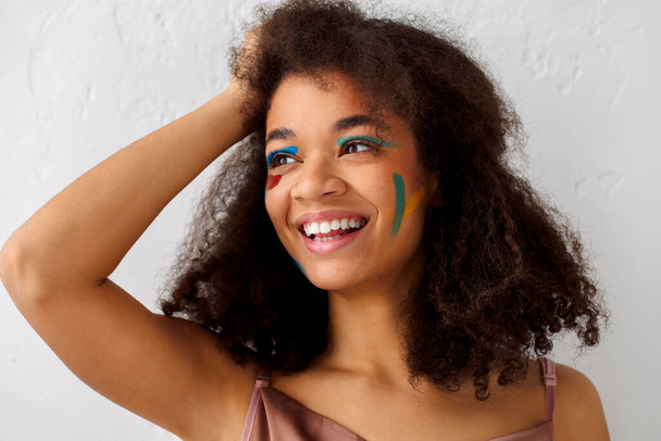Mulher de etnia afro-americana feliz bonita com maquiagem de arte criativa olhando de lado com sorriso agradável, menina encaracolada preta alegre com manchas de cor no rosto posando contra fundo de estúdio cinza - Foto, Imagem