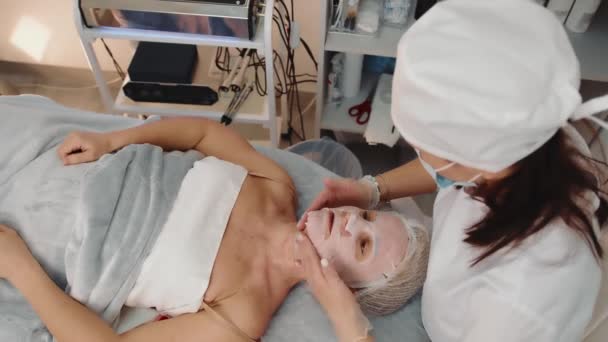 Vue latérale du cosmétologue en masque faisant un massage du cou à une patiente adulte dans un salon de beauté. Vue ci-dessus de la femme caucasienne recevant un massage du visage et du cou, relaxant sur le canapé. Concept de soins de beauté - Séquence, vidéo