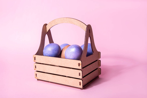 Wooden craft basket with purple painted shiny Easter eggs on a pink background. Easter holiday concept - Photo, Image