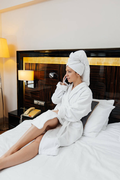 A young beautiful girl in a white coat is talking on the phone in her hotel room. Rest and travel. Hotel recreation, and tourism. - Photo, image