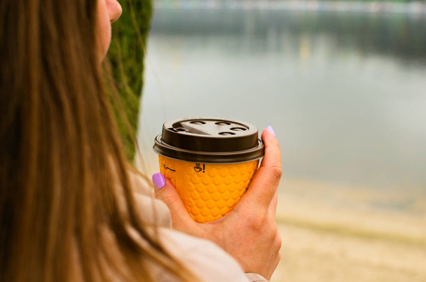 女性の手に黄色のコーヒーペーパーカップ。自然を背景にしている。大都市におけるコーヒーブレイクの概念. - 写真・画像