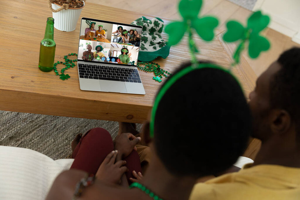 Африканская американская пара делает видео-звонок в день Святого Патрика друзьям на домашнем ноутбуке. празднование дня ирландского святого покровителя дома в изоляции во время карантинной изоляции. - Фото, изображение