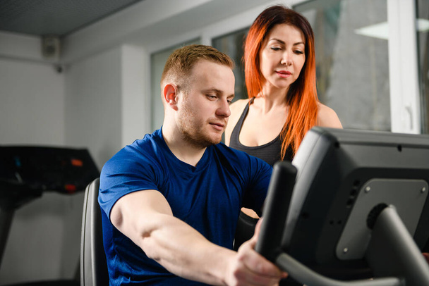 Женщина-тренер учит мужчину пользоваться оборудованием в современном тренажерном зале, смотреть на экран вместе, начинать тренировки - Фото, изображение