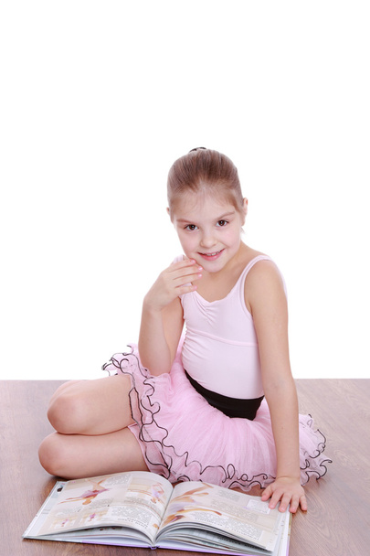 Studio image of a beautiful little ballerina in a pink tutu sitting on a hardwood floor for dancing and reading a book - Foto, Bild