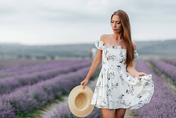 A young beautiful girl in a delicate dress and hat walks through a beautiful field of lavender and enjoys the fragrance of flowers. Rest and beautiful nature. Lavender blooming and flower picking. - Foto, Imagem