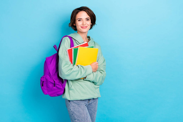 Photo of pretty shiny lady dressed green pullover backpack holding book stack empty space isolated blue color background - Foto, afbeelding