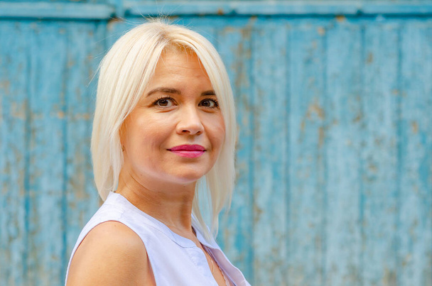 Gülümseyen, bakımlı orta yaşlı bir kadının portresi. Sarışın. Parlak makyaj. Bulanık mavi arkaplan - Fotoğraf, Görsel