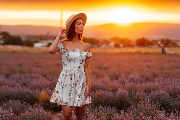 A young beautiful girl in a delicate dress and hat walks through a beautiful field of lavender and enjoys the fragrance of flowers. Rest and beautiful nature. Lavender blooming and flower picking. - Foto, afbeelding