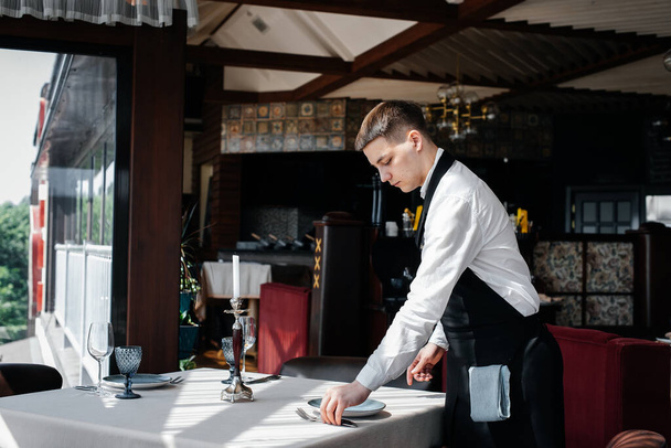 A young male waiter in a stylish uniform is engaged in serving the table in a beautiful gourmet restaurant. A high-level restaurant. Table service in the restaurant. - Foto, Imagem