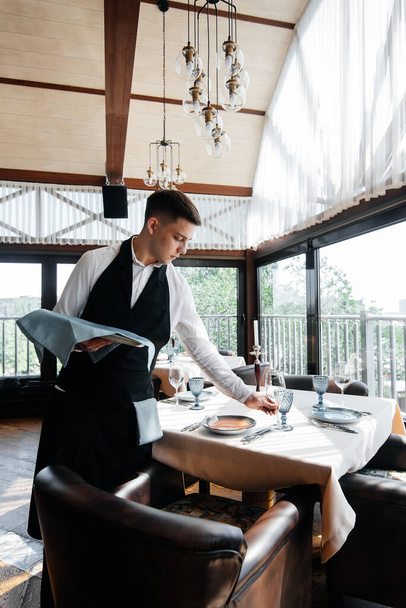 A young male waiter in a stylish uniform is engaged in serving the table in a beautiful gourmet restaurant. A high-level restaurant. Table service in the restaurant. - 写真・画像