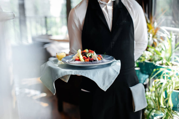 Крупним планом молодий офіціант у стильній формі несе вишуканий салат для клієнта в красивому ресторані гурманів. Обслуговування столів у ресторані
. - Фото, зображення