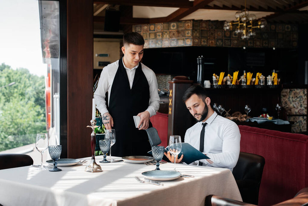 A young businessman in a fine restaurant examines the menu and makes an order to a young waiter in a stylish apron. Customer service. Table service in the restaurant. - Fotó, kép
