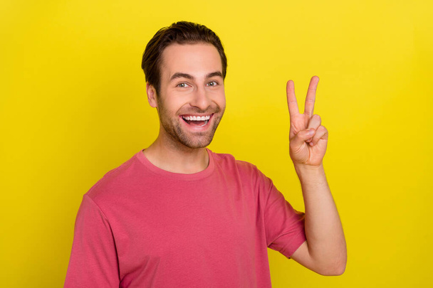 Photo of friendly positive guy show v-sign peaceful symbol wear pink t-shirt isolated yellow color background - Фото, изображение