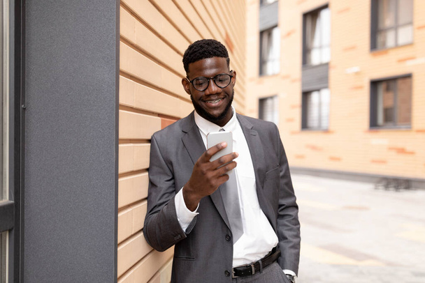 Concepto de estilo de vida. Elegante hombre de negocios afro navegar por Internet en el teléfono inteligente, apoyado en la pared del edificio de oficinas - Foto, imagen