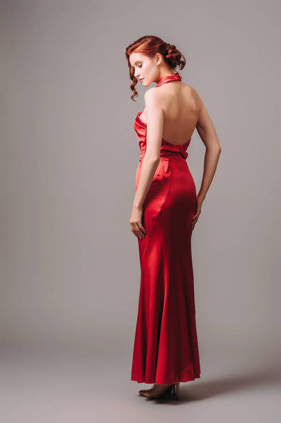 赤いノースリーブのドレスにギャザーとグレーのスタジオ背景に高いホルターネックラインで魅力的なセクシーな女性。ファッショナブルな女性の表情。自然の化粧やヘアドアのコンセプト. - 写真・画像