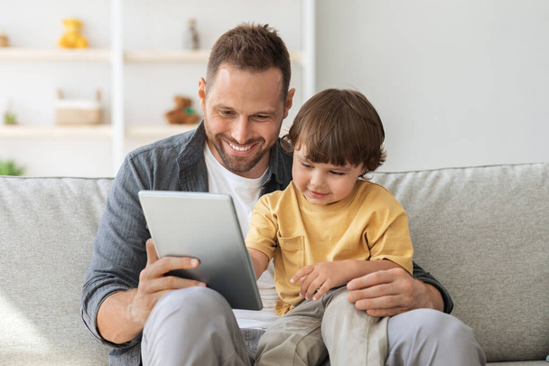 Lasten kasvatussovellus. Suloinen pieni poika pelaa kehitysvideopelejä digitaalisella tabletilla, istuu isän kanssa sohvalla - Valokuva, kuva