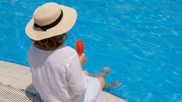 mulher em um chapéu de palha relaxa perto da piscina, bebe um coquetel rosa e balança as pernas na água. Conceito de humor verão - Foto, Imagem
