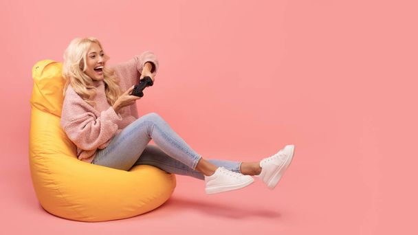 Joyful woman playing video games with joystick, sitting on beanbag chair over pink studio background, free space - Photo, image