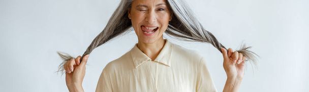 Middle aged Asian lady grimaces and holds long silver hair on light grey background - Foto, afbeelding