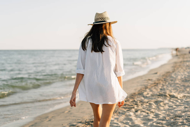 Το πίσω μέρος μιας γυναίκας με πουκάμισο και καπέλο περπατά σε μια ηλιόλουστη παραλία με τη γαλάζια θάλασσα στο βάθος. Η έννοια των διακοπών και των διακοπών. αντίγραφο χώρου - Φωτογραφία, εικόνα
