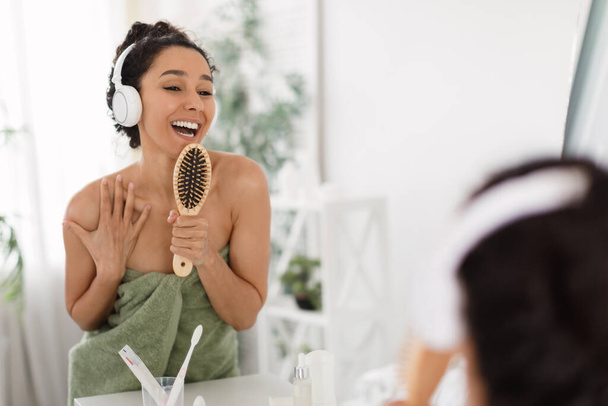 Ευτυχισμένη νεαρή γυναίκα με ακουστικά που απολαμβάνει μουσική, τραγούδι και χορό, χρησιμοποιώντας βούρτσα μαλλιών ως μικρόφωνο, διασκεδάζοντας κοντά στον καθρέφτη - Φωτογραφία, εικόνα