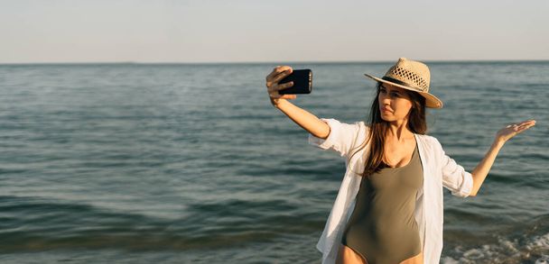 白い服の女性は、熱帯のビーチで休暇中の夕方に夏の日の日没に屋外の海のビーチの日の出に携帯電話のアプリケーションを使用してビデオ通話を行います。バナー. - 写真・画像