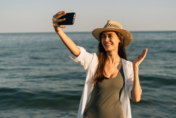 Close-up portrait of a girl on the ocean. Wearing a straw hat, she is holding a smartphone and making a video call using an app on her smartphone. accessible internet concept. Banner. copy space - Фото, зображення