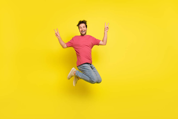 Full size photo of impressed brunet young guy jump show v-sign wear t-shirt jeans sneakers isolated on yellow background - Foto, immagini