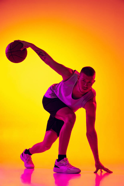Estúdio tiro de homem musculado, treinamento jogador de basquete com bola isolada em gradiente amarelo fundo roxo em luz de néon. Beleza, esporte, movimento, conceitos de atividade. - Foto, Imagem