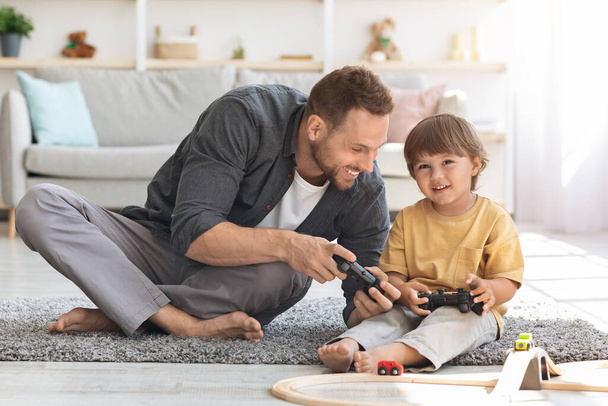 Молодий люблячий батько грає у відеоігри зі своїм милим маленьким сином, сидячи на підлозі з джойстиками, дитина дивиться на телевізор
 - Фото, зображення