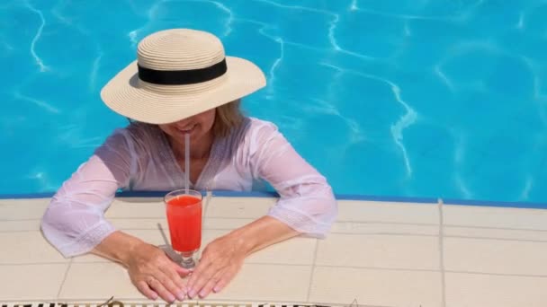 mujer rubia sonriente en sombrero de paja relaja y bebe cóctel naranja cerca de la piscina en un día soleado de verano. Concepto de humor de verano, vacaciones, todo incluido - Metraje, vídeo