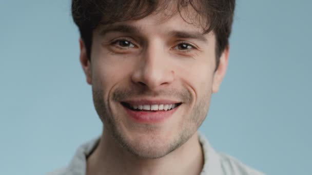close-up portret van jonge kalme knappe jongen met mooie ogen kijken naar de camera en glimlachen, blauwe studio achtergrond - Video