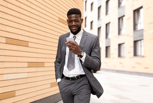Африканский американский генеральный директор, идущий рядом с офисным зданием, используя смартфон и улыбаясь, используя приложение или общаясь с клиентами - Фото, изображение