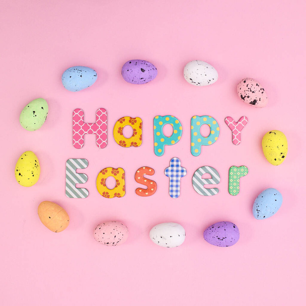 Creative spring holidays greeting card with Happy Easter text and colorful eggs around on pastel pink background. Flat lay - Fotó, kép