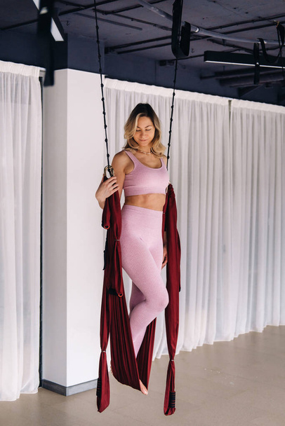 A woman in pink clothes does yoga on a suspended burgundy hammock in a bright gym. - Фото, зображення