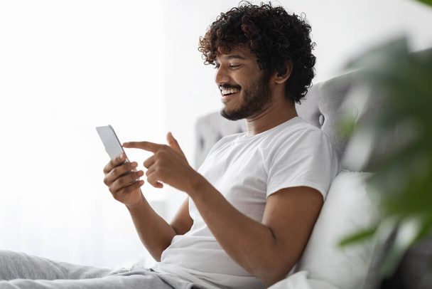 Positive dark-skinned guy scrolling in bed, using smartphone - Zdjęcie, obraz
