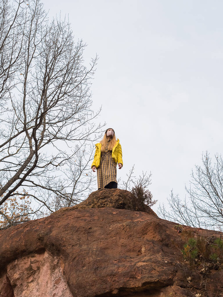 Женщина в маске для лица стоит на вершине скалы рядом с голыми деревьями на фоне облачного неба - Фото, изображение