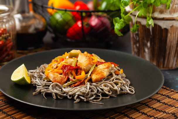 Wheat noodles with black sesame, fried in a wok with chicken and vegetables. Front view. Served on a black plate. - Zdjęcie, obraz