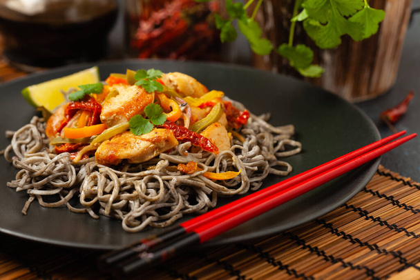 Wheat noodles with black sesame, fried in a wok with chicken and vegetables. Front view. Served on a black plate. - Foto, Imagem