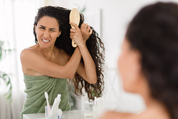 Frustrated young lady having problem brushing her tangled hair in front of mirror at home, copy space - Photo, Image